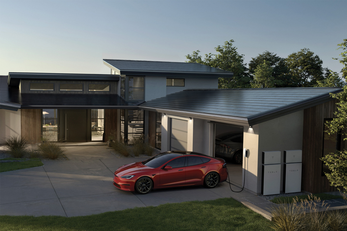 A modern home with Tesla solar roof shinlges, two Tesla Powerwall batteries, and a red Tesla electric car charging in the driveway