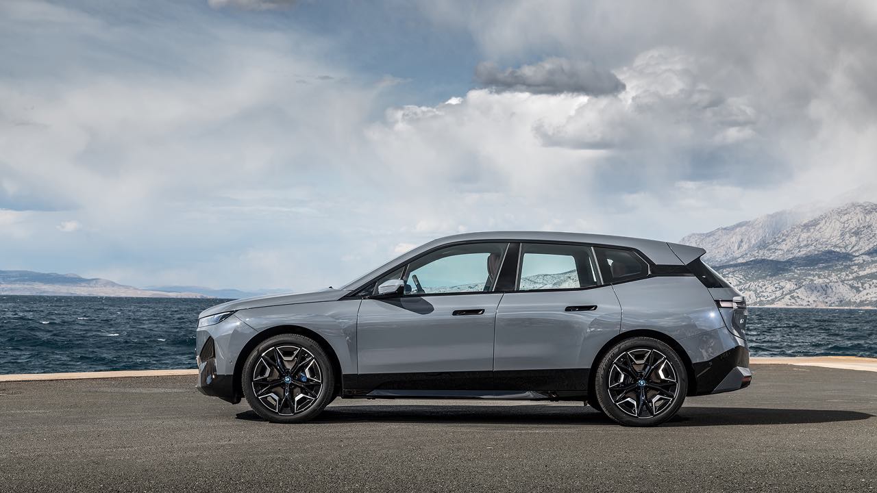 A grey BMW IX EV parked by a body of water with mountains in the background