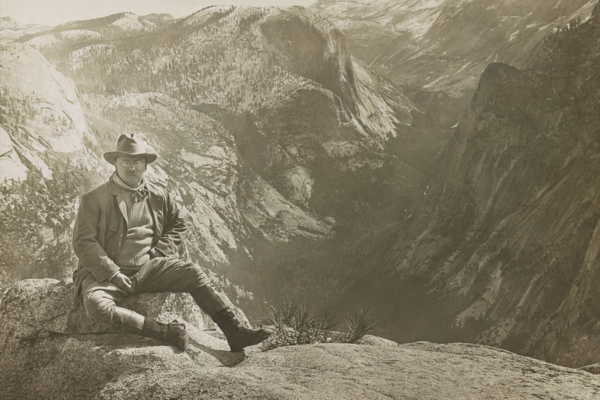 Theodore Roosevelt wearing outdoor gear sitting on a rock with mountains in the background