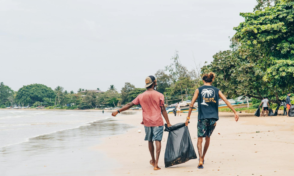 Two people holding a full black trash bag walking down a shoreline.